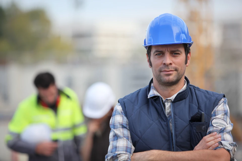 Chantier de construction en extérieur avec ingénieur ou chef de projet au premier plan portant casque de sécurité bleu et veste matelassée marine sur chemise à carreaux